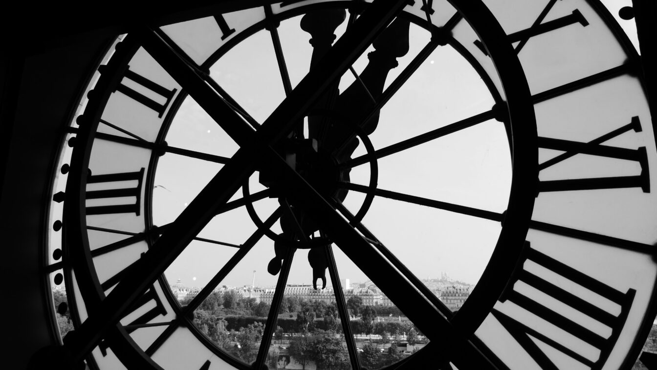 Blick durch eine große, historische Uhr mit römischen Ziffern, die von innen fotografiert wurde. Im Hintergrund ist eine verschwommene Stadtlandschaft zu sehen, möglicherweise durch das Glas verzerrt. Das Schwarz-Weiß-Foto betont die Details der Uhr und die Silhouette der dahinterliegenden Gebäude.