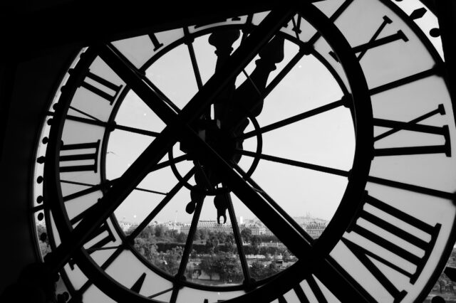 Blick durch eine große, historische Uhr mit römischen Ziffern, die von innen fotografiert wurde. Im Hintergrund ist eine verschwommene Stadtlandschaft zu sehen, möglicherweise durch das Glas verzerrt. Das Schwarz-Weiß-Foto betont die Details der Uhr und die Silhouette der dahinterliegenden Gebäude.