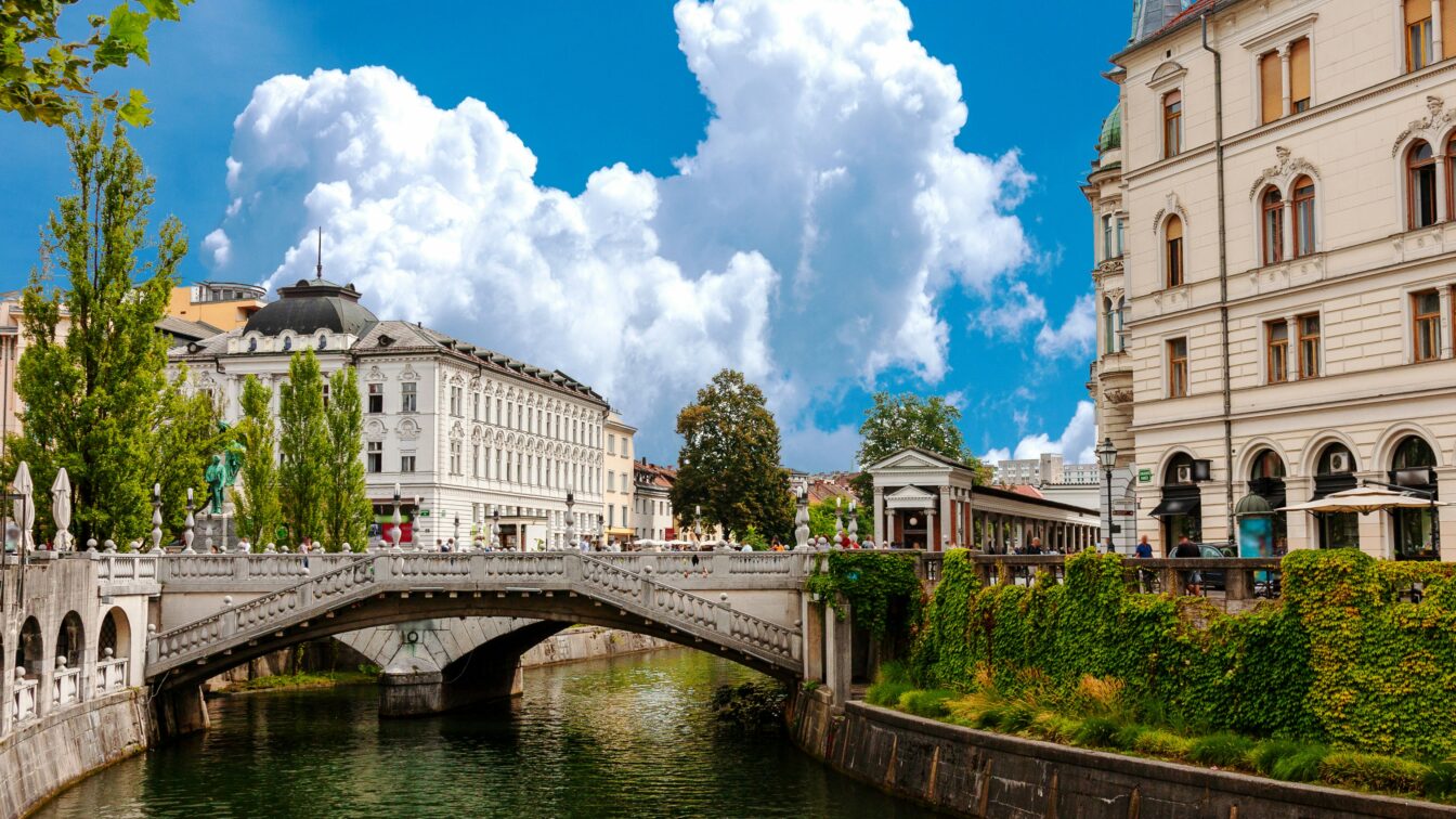 (c) Eugene Kuznetsov | Unsplash Stadtansicht von Ljubljana, Gebäude neben einem Fluss, große weiße Wolke