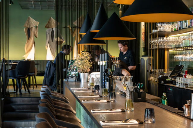 Stilvoll eingerichteter Innenraum des Restaurants Vina in Graz. Im Vordergrund eine elegante Bar mit gedeckten Plätzen, Flaschen und Gläsern. Schwarze, konisch geformte Lampen hängen über der Theke. Im Hintergrund bedienen Mitarbeitende an der Bar, während Gäste an Tischen im erhöhten Sitzbereich sitzen. Die grüne Wand mit großem Wandgemälde zweier stilisierter Figuren in traditionellen vietnamesischen Kleidern verleiht dem Raum ein besonderes Ambiente.