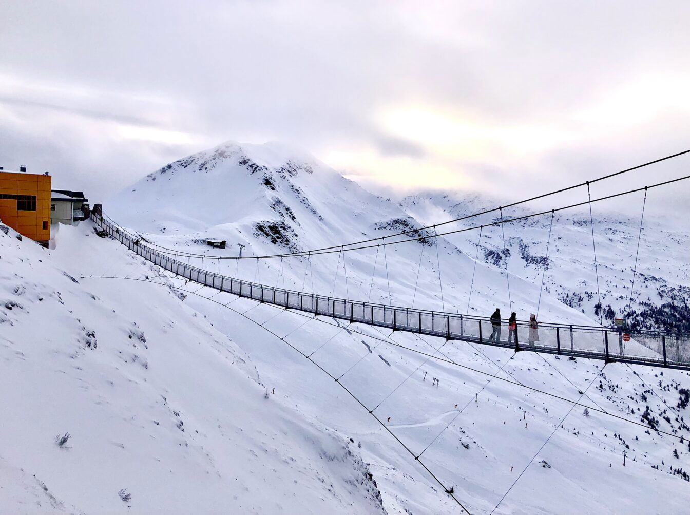 Eine Hängebrücke spannt sich über eine schneebedeckte, alpine Landschaft in den Bergen von Gastein. Drei Menschen gehen über die Brücke, die von stabilen Seilen gehalten wird. Im Hintergrund sind hohe, verschneite Berggipfel und ein wolkenverhangener Himmel zu sehen, der die Szene in kühles Licht taucht. Rechts neben der Brücke führt ein Skilift den Hang hinauf. Die Stimmung ist ruhig und winterlich.