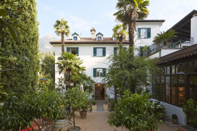 Das Ottmanngut, ein charmantes historisches Gebäude im mediterranen Stil, umgeben von Palmen und üppiger Vegetation. Im Vordergrund stehen Gartenmöbel und Töpfe mit Orangenbäumen, die eine einladende, ruhige Atmosphäre schaffen. Im Hintergrund sind Berge und blauer Himmel zu sehen.