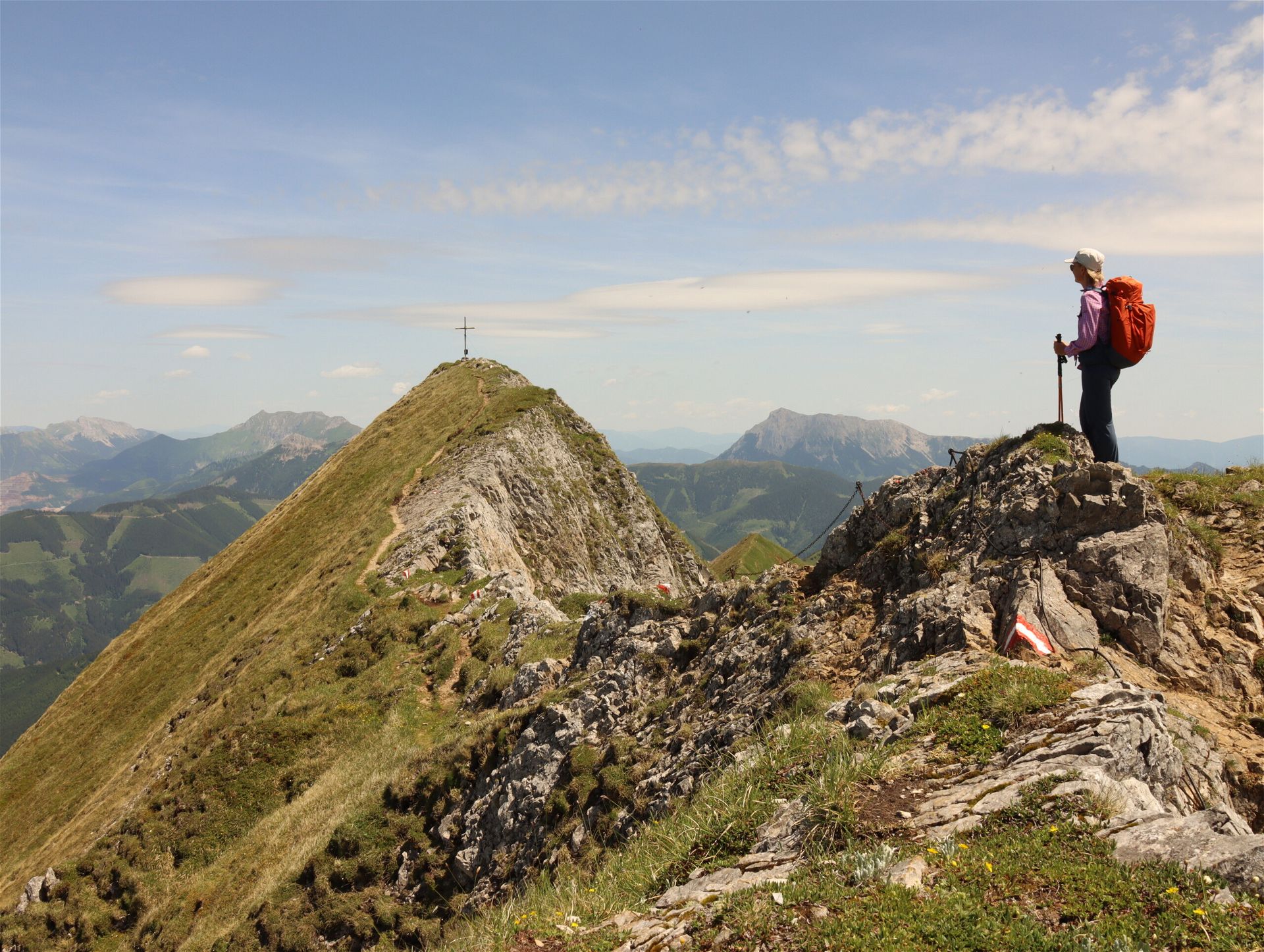 Eine Person steht am Gipfel eines Berges im Sommer bei Sonnenschein, dahinter sieht man eine weite Berglandschaft.