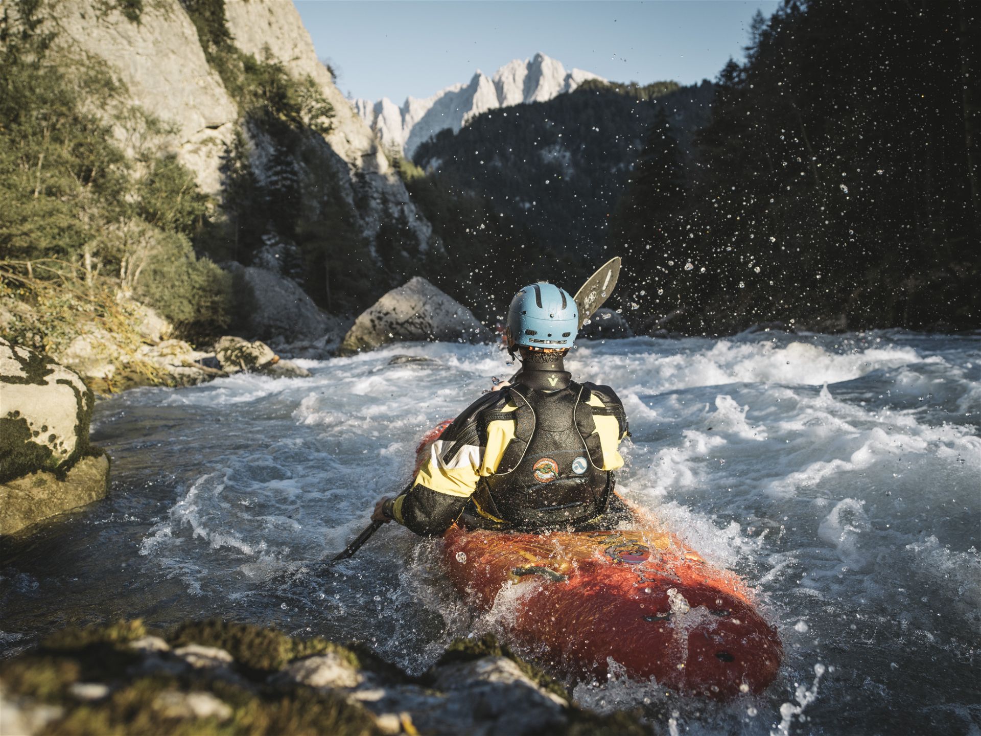 Auf dem Bild ist ein Kajak mit einer Person darin auf einem wilden Fluss zu sehen, mit Helm und Paddel inmitten einer schönen, grünen Naturkulisse.
