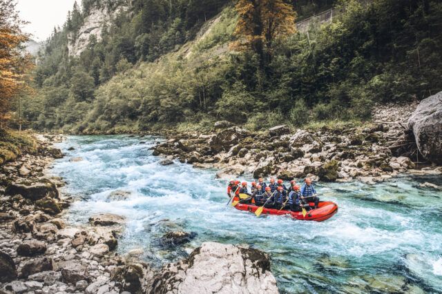 Auf dem Bild zu sehen ist ein Rafting-Boot auf einem wilden Fluss, darauf einige Menschen mit Helmen und Paddeln inmitten einer schönen, grünen Naturkulisse.
