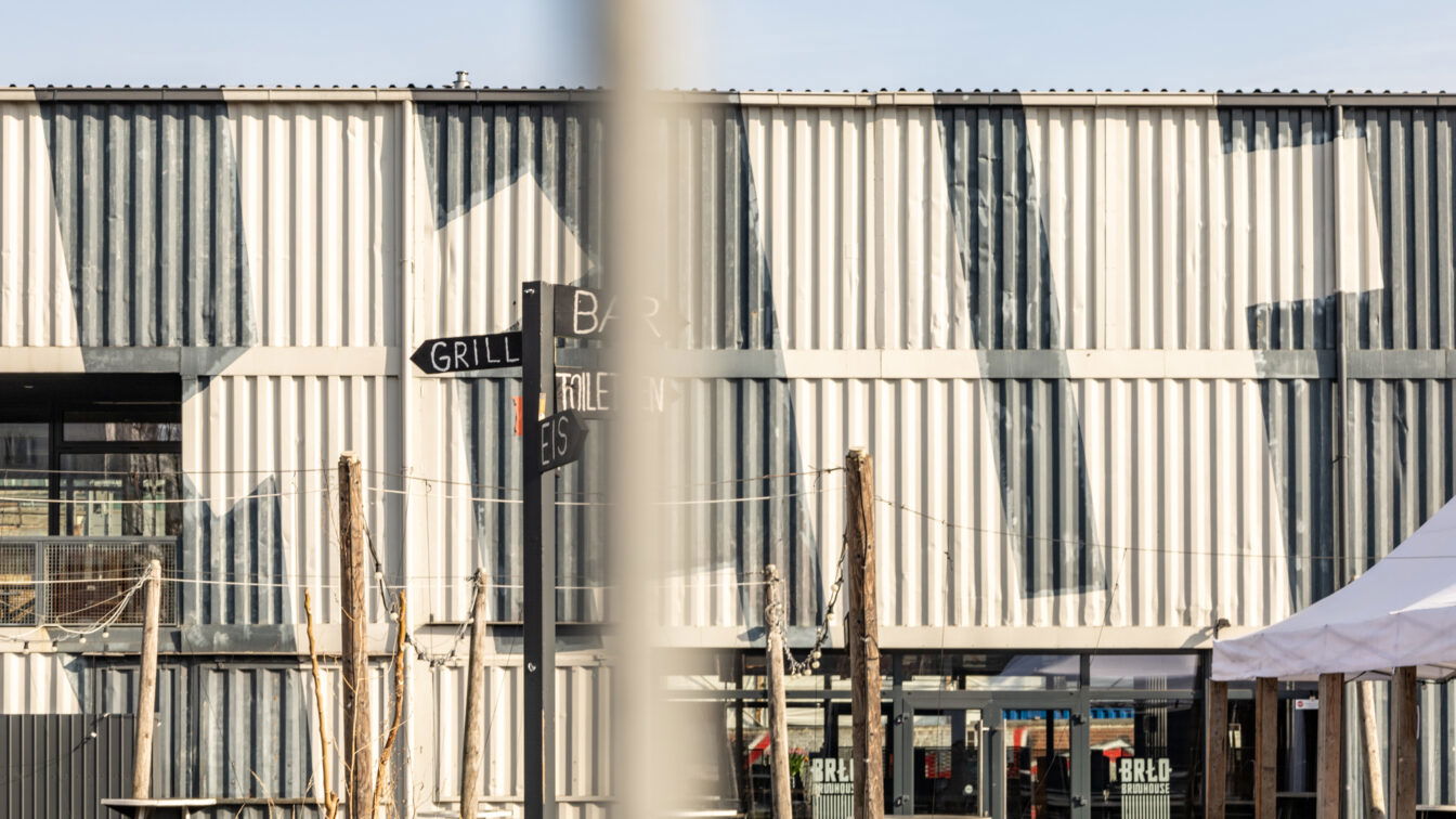 (c) Sonja Koller | 1000things Außenansicht des BRLO Brwhouse Biergartens in Berlin mit einer modernen, industriellen Ästhetik. Die Fassade besteht aus weiß-grau gestrichenen Schiffscontainern mit geometrischen Mustern. Im Vordergrund steht ein schwarzer Wegweiser mit den Beschriftungen 'Grill', 'Bar', 'Eis' und 'Toiletten'. Mehrere Holztische und Sitzgelegenheiten sind über das Gelände verteilt, während Lichterketten zwischen Holzpfosten gespannt sind. Auf der rechten Seite ist ein weißes Zelt zu sehen, das zum Außenbereich des Biergartens gehört. Die Szene wird von hellem Sonnenlicht beleuchtet, das starke Schatten auf die Wand wirft.