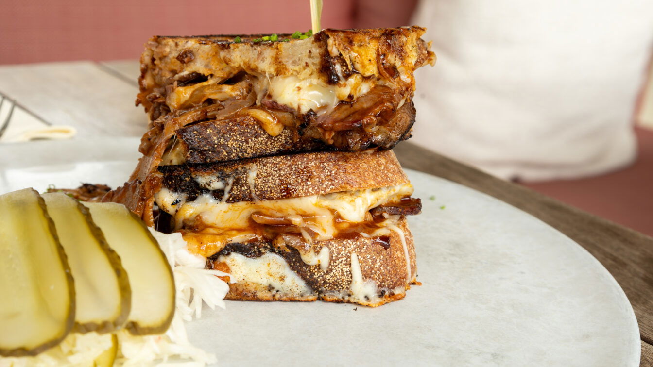 (c) Sonja Koller | 1000things Nahaufnahme eines herzhaften Grilled Cheese Sandwiches mit geschmolzenem Käse und Pulled Pork, serviert auf einem hellen Teller mit Sauerkraut und Gurkenscheiben. Das Gericht steht auf einem Holztisch in gemütlicher Restaurantatmosphäre in Budapest. Perfekt für Foodies auf der Suche nach den besten Bars und Restaurants in Budapest mit moderner, kreativer Küche und Wohlfühlfaktor.