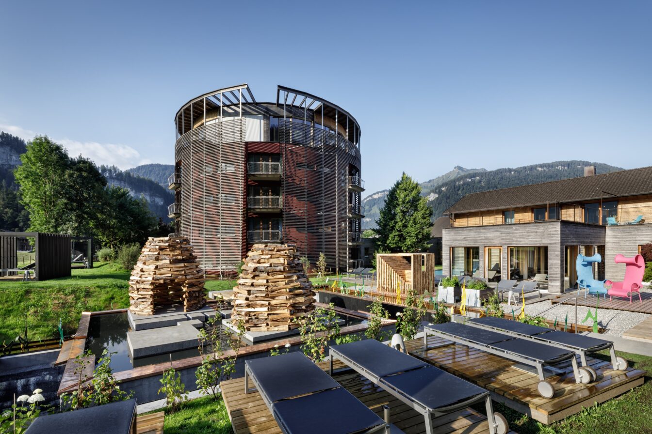 GAMS zu zweit, Hotels, Österreich, Außenansicht eines modernen, luxuriösen Hotelkomplexes, umgeben von malerischen Bergen und grüner Landschaft. Im Vordergrund sind Liegestühle auf Holzplattformen und zwei große, kunstvoll gestapelte Holzhaufen zu sehen. Der Hauptbau des Hotels hat eine runde Form mit mehreren Balkonen und einer einzigartigen Fassadenstruktur. Neben dem Hauptgebäude steht ein weiteres modernes Gebäude mit großen Fenstern, durch die der Innenraum sichtbar ist.