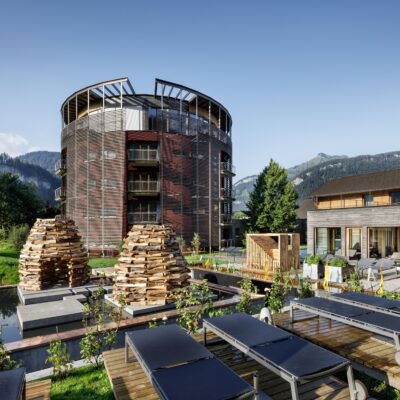 GAMS zu zweit, Hotels, Österreich, Außenansicht eines modernen, luxuriösen Hotelkomplexes, umgeben von malerischen Bergen und grüner Landschaft. Im Vordergrund sind Liegestühle auf Holzplattformen und zwei große, kunstvoll gestapelte Holzhaufen zu sehen. Der Hauptbau des Hotels hat eine runde Form mit mehreren Balkonen und einer einzigartigen Fassadenstruktur. Neben dem Hauptgebäude steht ein weiteres modernes Gebäude mit großen Fenstern, durch die der Innenraum sichtbar ist.