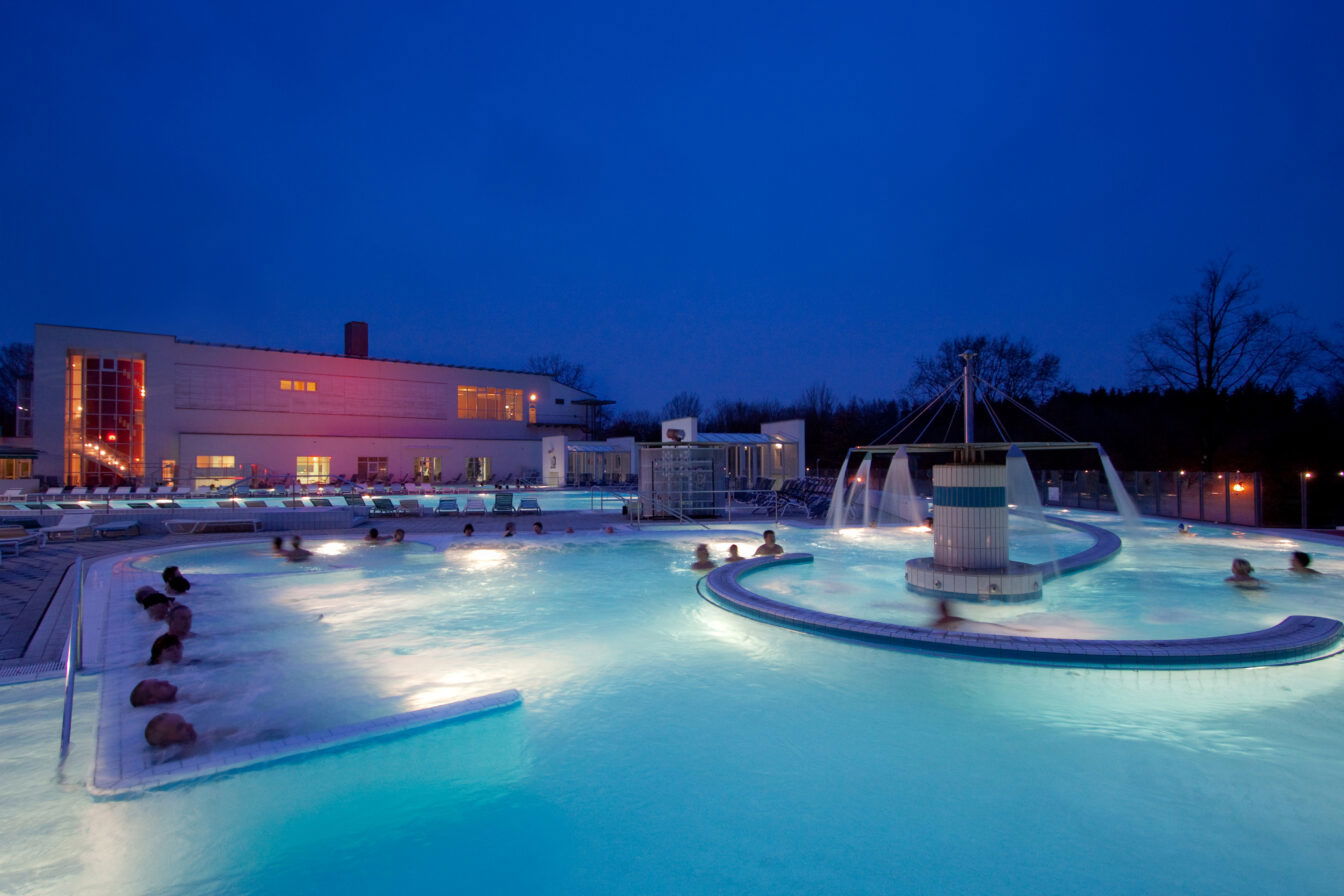 Das Bild zeigt einen Außenbereich der Europa Therme in Bad Füssing, Bayern, bei Nacht. Verschiedene Thermalpools sind beleuchtet, während Badegäste entspannen. Im Vordergrund sieht man einen kreisförmigen Pool mit Wasserdüsen, die sanft sprudeln, während Menschen im warmen Wasser sitzen. Im Hintergrund ist das Hauptgebäude der Therme zu sehen, das ebenfalls in stimmungsvollem Licht erstrahlt.