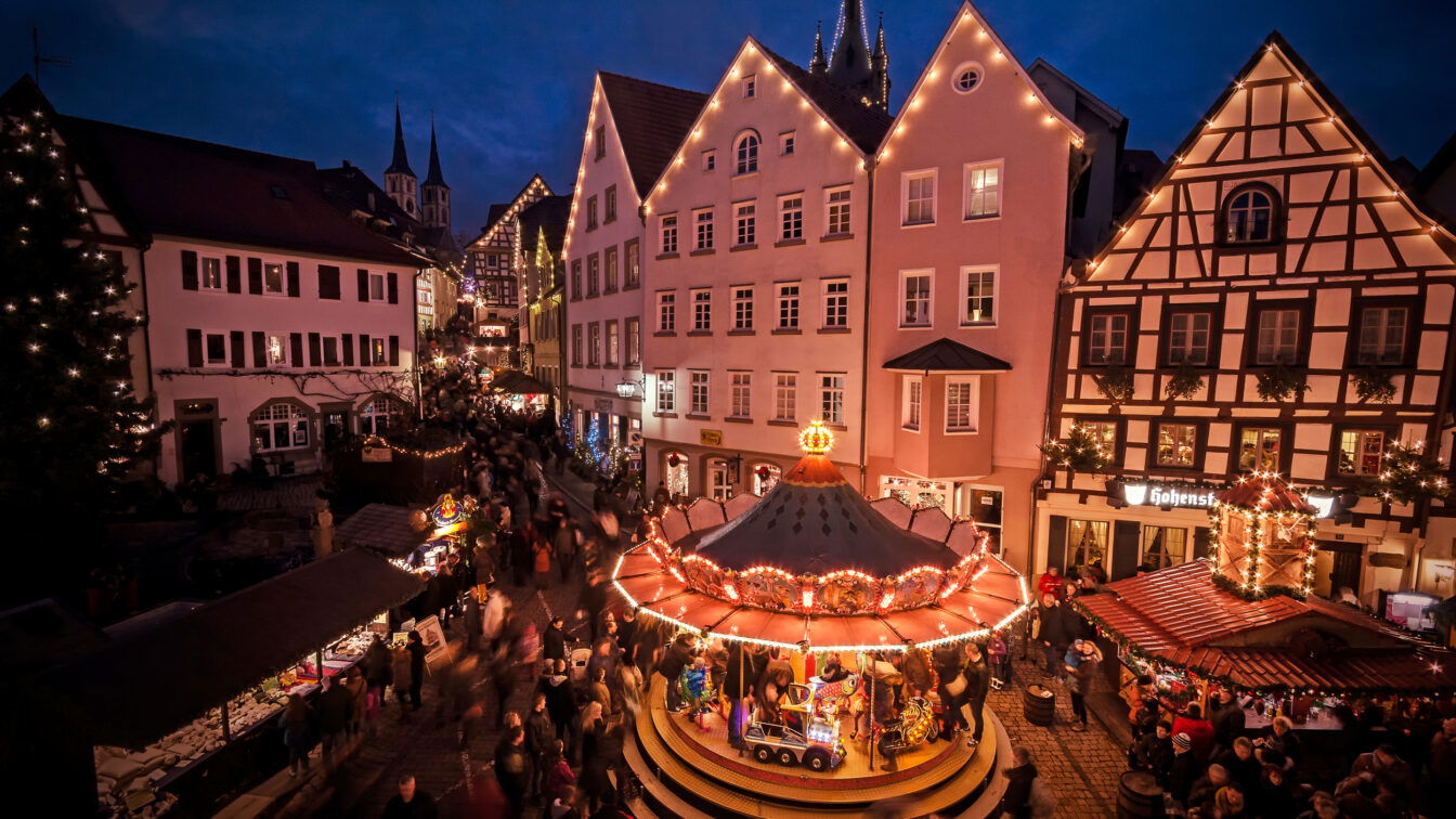 Das Bild zeigt einen festlich beleuchteten Weihnachtsmarkt in einer historischen Altstadt am Abend. Im Vordergrund befindet sich ein traditionelles Kinderkarussell mit bunten Lichtern und mehreren Figuren, darunter ein Zug und Pferde. Rund um den Platz sind dekorierte Fachwerkhäuser und weitere Stände des Weihnachtsmarkts zu sehen, die von Menschen besucht werden. Im Hintergrund ragen Kirchtürme in den Abendhimmel, während die Atmosphäre von Lichtern und weihnachtlicher Stimmung geprägt ist