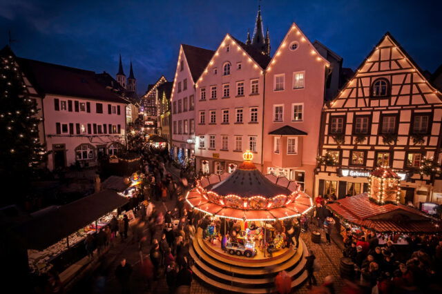 Das Bild zeigt einen festlich beleuchteten Weihnachtsmarkt in einer historischen Altstadt am Abend. Im Vordergrund befindet sich ein traditionelles Kinderkarussell mit bunten Lichtern und mehreren Figuren, darunter ein Zug und Pferde. Rund um den Platz sind dekorierte Fachwerkhäuser und weitere Stände des Weihnachtsmarkts zu sehen, die von Menschen besucht werden. Im Hintergrund ragen Kirchtürme in den Abendhimmel, während die Atmosphäre von Lichtern und weihnachtlicher Stimmung geprägt ist