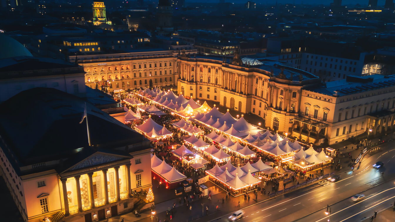 Ein festlich beleuchteter Weihnachtsmarkt am Bebelplatz in Berlin bei Nacht. Reihenweise weiße Zelte mit goldenen Lichtern, die den Platz in ein warmes, gemütliches Licht tauchen, während Menschen zwischen den Ständen flanieren. Die historische Architektur des Bebelplatzes mit imposanten Gebäuden im Hintergrund verstärkt die festliche Atmosphäre. Vor dem Markt befindet sich eine Straße mit vorbeifahrenden Autos.