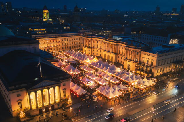 Ein festlich beleuchteter Weihnachtsmarkt am Bebelplatz in Berlin bei Nacht. Reihenweise weiße Zelte mit goldenen Lichtern, die den Platz in ein warmes, gemütliches Licht tauchen, während Menschen zwischen den Ständen flanieren. Die historische Architektur des Bebelplatzes mit imposanten Gebäuden im Hintergrund verstärkt die festliche Atmosphäre. Vor dem Markt befindet sich eine Straße mit vorbeifahrenden Autos.