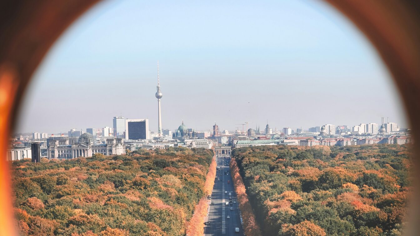 Das Bild zeigt einen Blick auf Berlin im Herbst, aufgenommen durch einen runden Rahmen. Im Zentrum verläuft eine lange, gerade Straße, die durch einen großen Park mit herbstlich gefärbten Bäumen führt. Im Hintergrund ragt der Berliner Fernsehturm markant aus der Skyline hervor, umgeben von weiteren Gebäuden wie dem Reichstagsgebäude.