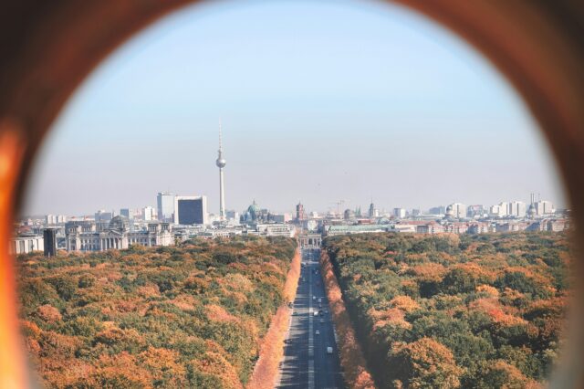 Das Bild zeigt einen Blick auf Berlin im Herbst, aufgenommen durch einen runden Rahmen. Im Zentrum verläuft eine lange, gerade Straße, die durch einen großen Park mit herbstlich gefärbten Bäumen führt. Im Hintergrund ragt der Berliner Fernsehturm markant aus der Skyline hervor, umgeben von weiteren Gebäuden wie dem Reichstagsgebäude.