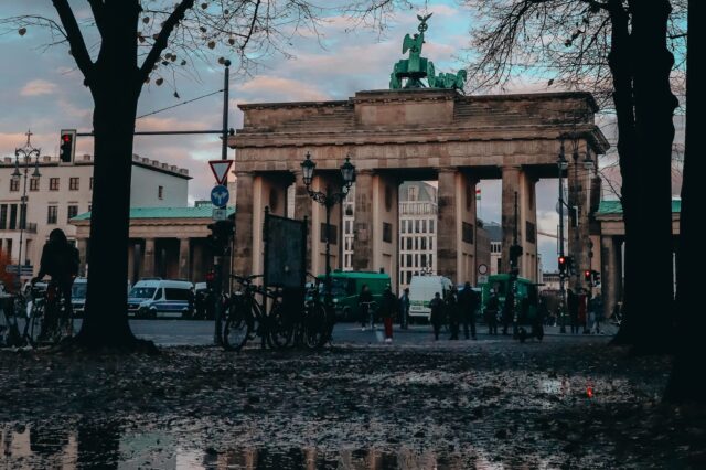 Blick auf das Brandenburger Tor in Berlin an einem bewölkten Tag. Im Vordergrund spiegeln sich Bäume und das Tor in einer Wasserpfütze. Menschen und Fahrräder sind um das Tor herum zu sehen, während die Lichter der Ampeln leuchten.