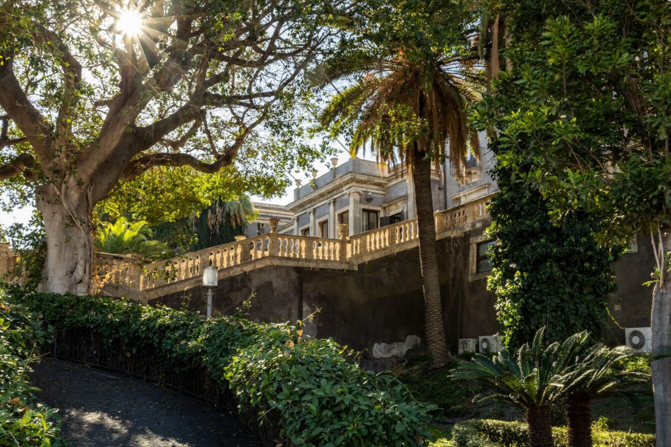 Ein prachtvolles historisches Gebäude in Catania, umgeben von üppigem Grün und Palmen. Sonnenstrahlen brechen durch die Bäume, was der Szene eine friedliche Atmosphäre verleiht.