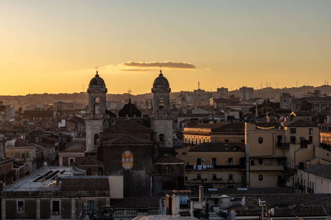 Ein Blick auf Catania, Italien, bei Sonnenuntergang. Die Silhouette zweier Kirchtürme dominiert den Vordergrund, während die Stadt im warmen Abendlicht leuchtet.