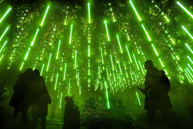 Menschen stehen unter einem dichten, leuchtenden Netz aus vertikal hängenden grünen Lichtstäben und funkelnden LED-Lichtern, die eine mystische, futuristische Atmosphäre schaffen. Die Silhouetten der Personen heben sich dunkel vom intensiven grünen Licht im Hintergrund ab.