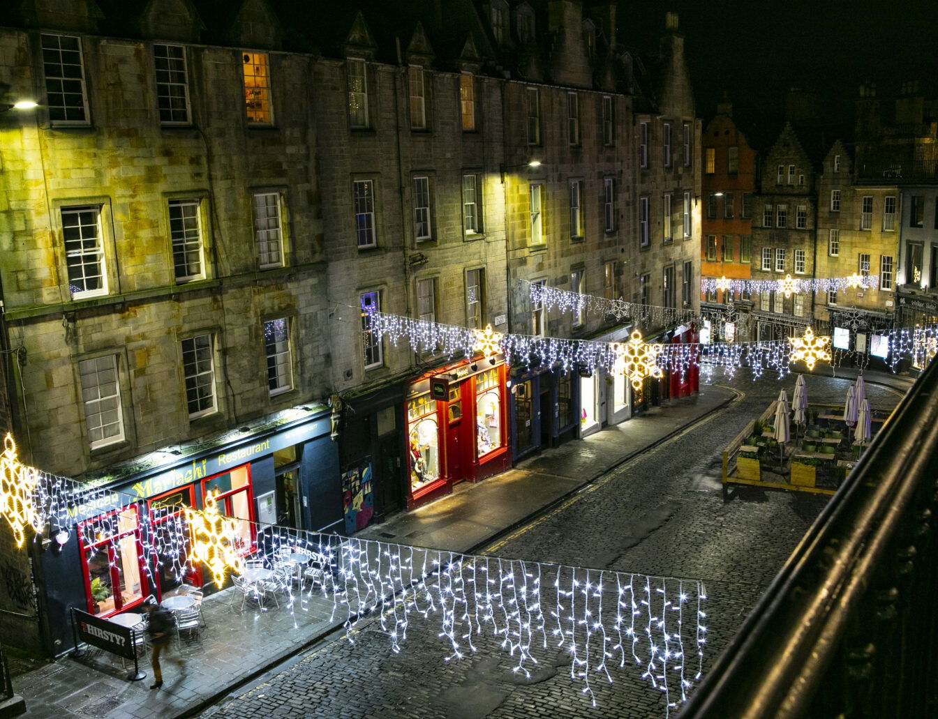 Ein festlich beleuchteter Straßenabschnitt in Edinburgh bei Nacht. Die historischen Gebäude auf der Victoria Street sind von Lichterketten und leuchtenden Sternen dekoriert, die über die Straße gespannt sind und eine gemütliche, weihnachtliche Atmosphäre schaffen.