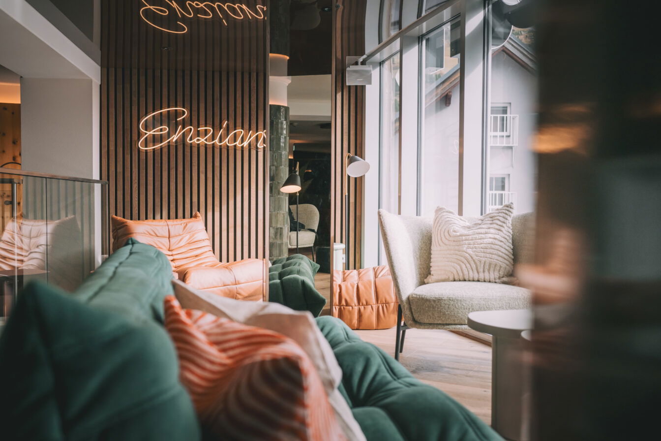 Enzian, Hotel, Adult Only, Ein modernes, gemütliches Hotel-Interieur mit eleganten Holzwänden und einer Neon-Leuchtschrift "Enzian". Weiche, grüne und orangefarbene Sofas sind um einen hellen Raum verteilt, der von großen Fenstern mit viel Tageslicht durchflutet wird. Ein bequemer, grauer Sessel mit weißen, strukturierten Kissen steht neben einem kleinen runden Tisch. Das Ambiente wirkt einladend und entspannend.