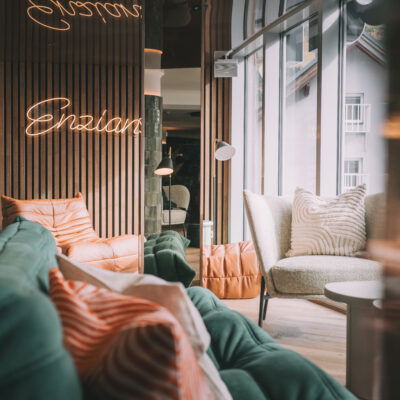 Enzian, Hotel, Adult Only, Ein modernes, gemütliches Hotel-Interieur mit eleganten Holzwänden und einer Neon-Leuchtschrift "Enzian". Weiche, grüne und orangefarbene Sofas sind um einen hellen Raum verteilt, der von großen Fenstern mit viel Tageslicht durchflutet wird. Ein bequemer, grauer Sessel mit weißen, strukturierten Kissen steht neben einem kleinen runden Tisch. Das Ambiente wirkt einladend und entspannend.