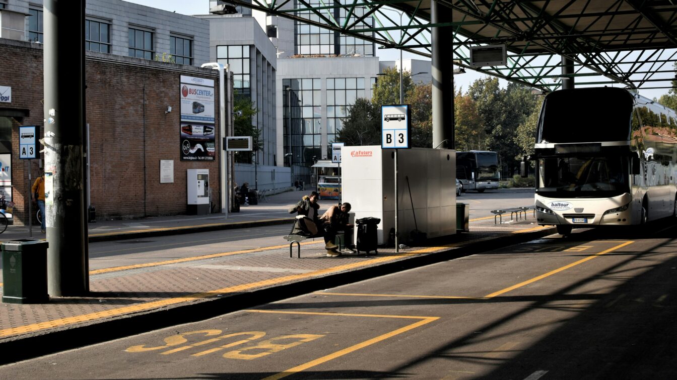 Das Bild zeigt eine Busstation an einem sonnigen Tag. Im Vordergrund befindet sich ein leerer Busparkplatz, auf dem der Schriftzug "BUS" gelb auf den Asphalt gemalt ist. Rechts steht ein großer weißer Reisebus mit einem Schriftzug "baltour" an der Front. Daneben sitzen zwei Personen auf einer Bank unter einer Überdachung und warten. Hinter ihnen befinden sich moderne Gebäude mit großen Glasfenstern. Die Szenerie wirkt ruhig, es gibt keine sichtbare Hektik oder Menschenmenge. Schilder mit Busnummern (z.B. B3) kennzeichnen die Abfahrtsbereiche.