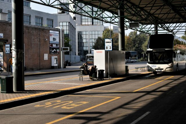 Das Bild zeigt eine Busstation an einem sonnigen Tag. Im Vordergrund befindet sich ein leerer Busparkplatz, auf dem der Schriftzug "BUS" gelb auf den Asphalt gemalt ist. Rechts steht ein großer weißer Reisebus mit einem Schriftzug "baltour" an der Front. Daneben sitzen zwei Personen auf einer Bank unter einer Überdachung und warten. Hinter ihnen befinden sich moderne Gebäude mit großen Glasfenstern. Die Szenerie wirkt ruhig, es gibt keine sichtbare Hektik oder Menschenmenge. Schilder mit Busnummern (z.B. B3) kennzeichnen die Abfahrtsbereiche.