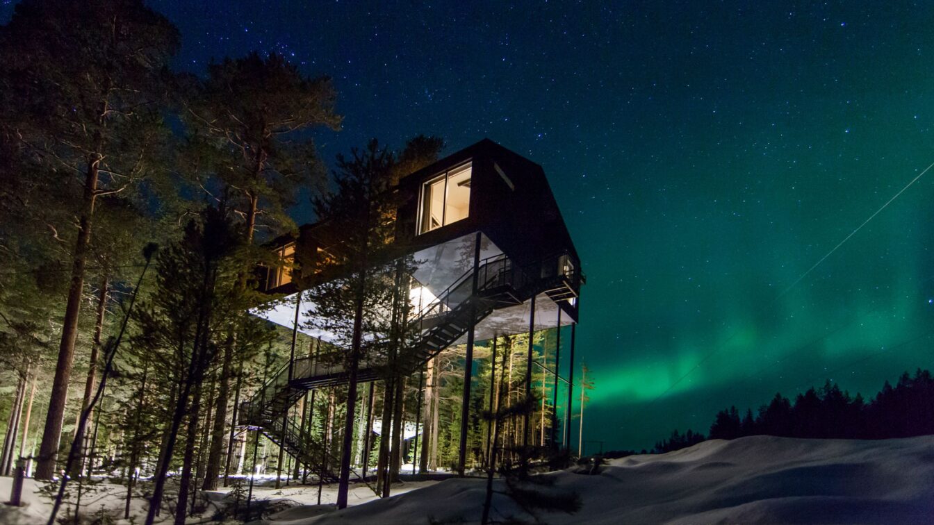 Das Bild zeigt ein modernes Baumhaus-Hotel, das hoch auf Stelzen inmitten eines winterlichen Waldes in Schweden steht. Der Nachthimmel ist mit Sternen bedeckt und die Nordlichter leuchten in intensiven Grüntönen. Eine Außentreppe führt zum erhöhten Eingang des Gebäudes, das teilweise von Bäumen umgeben ist. Die Szenerie vermittelt eine ruhige, abgelegene Atmosphäre in der Natur.