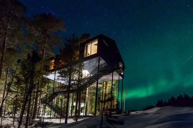 Das Bild zeigt ein modernes Baumhaus-Hotel, das hoch auf Stelzen inmitten eines winterlichen Waldes in Schweden steht. Der Nachthimmel ist mit Sternen bedeckt und die Nordlichter leuchten in intensiven Grüntönen. Eine Außentreppe führt zum erhöhten Eingang des Gebäudes, das teilweise von Bäumen umgeben ist. Die Szenerie vermittelt eine ruhige, abgelegene Atmosphäre in der Natur.