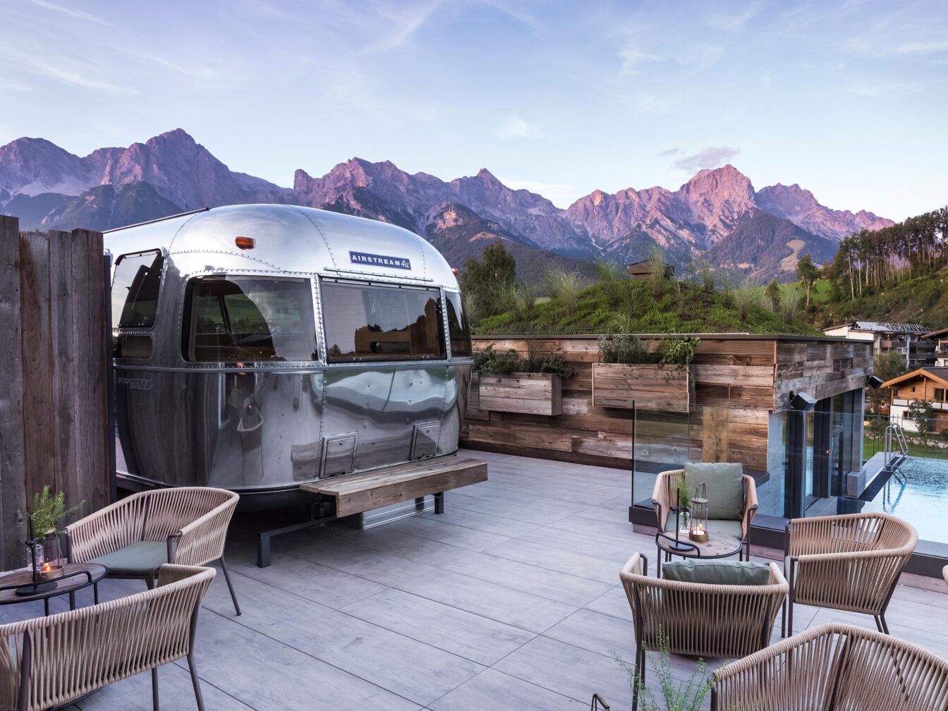 Hotel Sepp, Eine stilvolle Terrasse mit einem glänzenden Airstream-Wohnwagen, umgeben von gemütlichen Sitzmöbeln. Im Hintergrund erstrecken sich majestätische Berge unter einem klaren Himmel im warmen Licht der untergehenden Sonne. Neben dem Wohnwagen befindet sich ein Infinity-Pool mit Glasgeländer, das den Blick auf die umliegende Natur freigibt. Die Dachterrasse ist mit Pflanzen dekoriert, und das Ambiente vermittelt eine entspannte, luxuriöse Atmosphäre