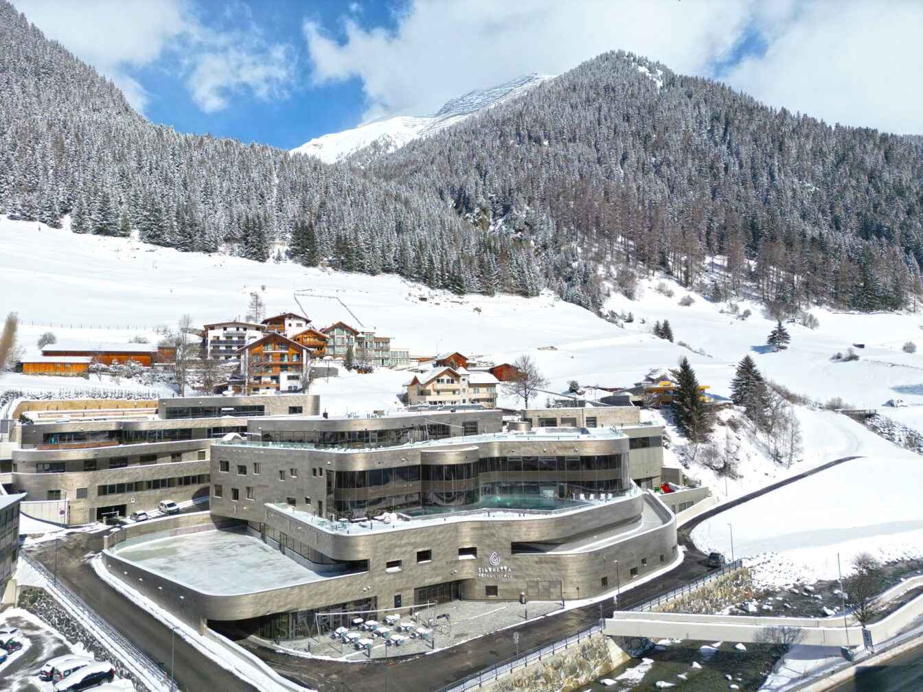 Luftaufnahme der Silvretta Therme in Ischgl im Winter. Das moderne Gebäude der Therme ist in einer kurvigen, futuristischen Architektur gestaltet und besteht aus glatten, grauen Fassaden. Umgeben ist die Therme von schneebedeckten Bergen und Wäldern, die das Bild einer typischen alpinen Winterlandschaft vermitteln