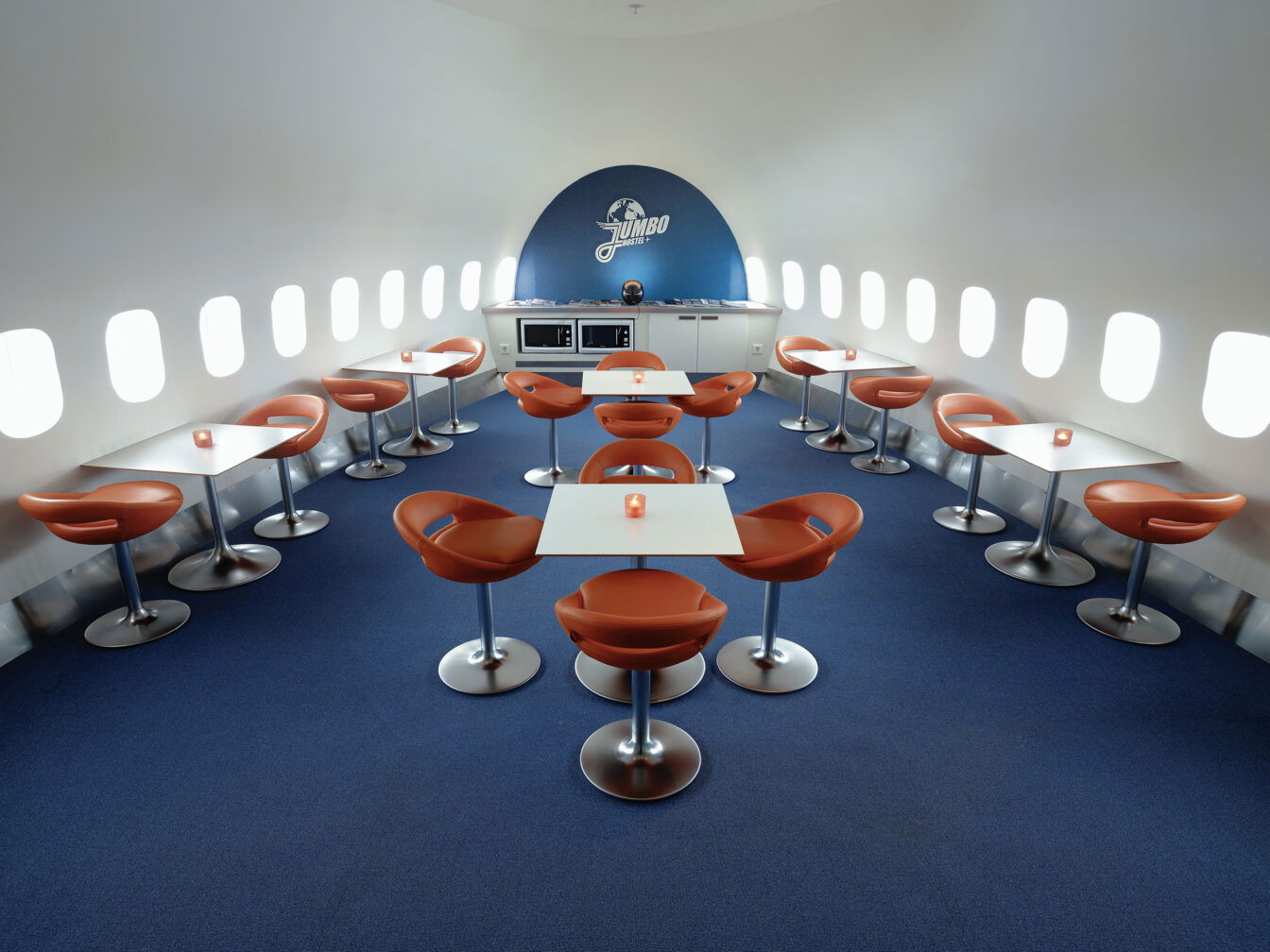 Das Bild zeigt den Innenraum eines Cafés oder Aufenthaltsraums im JumboStay Hotel, einem umgebauten Flugzeug am Flughafen Stockholm-Arlanda. Die Tische und Stühle sind in zwei Reihen angeordnet, mit futuristischen, orangefarbenen Stühlen und weißen Tischen. Der Boden ist mit einem blauen Teppich ausgelegt, während die Wand mit Flugzeugfenstern durchzogen ist, was den Flugzeugcharakter des Raums betont. Im hinteren Bereich des Raumes ist eine kleine Küchenzeile mit Mikrowellen zu sehen. Über dieser hängt ein Logo mit der Aufschrift „Jumbo“. Die Einrichtung kombiniert eine moderne und minimalistische Atmosphäre mit einem einzigartigen Flugzeugdesign.