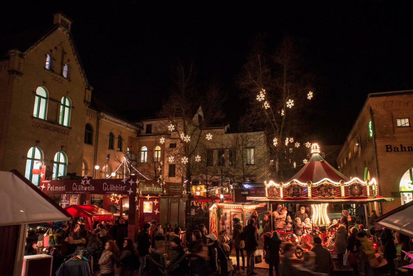 Ein stimmungsvoller Weihnachtsmarkt auf dem Gelände der Kulturbrauerei in Berlin bei Nacht. Im Vordergrund sind verschiedene Stände mit Schildern für Glühwein und Pelmeni zu sehen, umgeben von Menschen, die durch den Markt schlendern. Ein nostalgisches Karussell mit leuchtenden Lichtern zieht Kinder und Familien an. Über den Bäumen hängen leuchtende Schneeflocken als Dekoration, die die Atmosphäre warm und einladend wirken lassen. Die alten Backsteingebäude der Kulturbrauerei bilden den Hintergrund und verleihen dem Markt einen charmanten, historischen Rahmen