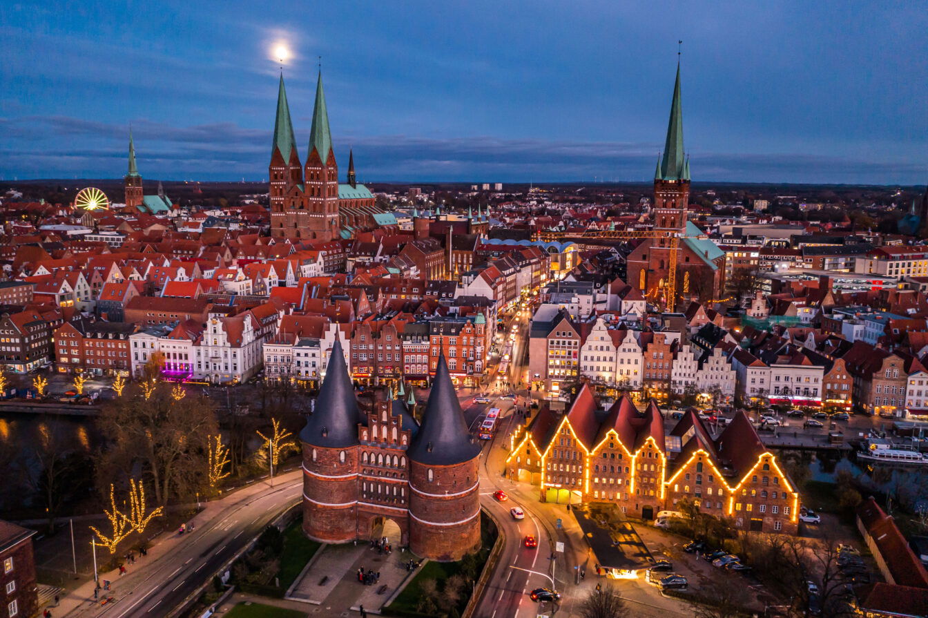 Das Bild zeigt eine Panoramaaufnahme der Altstadt von Lübeck in der Dämmerung. Die Stadt ist festlich beleuchtet, mit Lichterketten, die die Gebäude und Bäume schmücken. Im Vordergrund ist das beleuchtete Holstentor zu sehen, während im Hintergrund die Türme der Marienkirche und des Doms von Lübeck emporragen. Die Straßen und Plätze der Stadt sind von Weihnachtsmarktständen und geschmückten Gebäuden umgeben, was eine festliche Atmosphäre schafft.