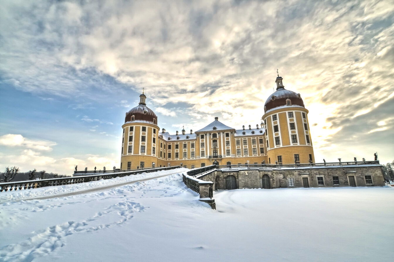 Das Bild zeigt das Schloss Moritzburg im Winter. Der Barockbau hebt sich mit seiner gelben Fassade und den vier markanten Rundtürmen vor einem bewölkten Himmel ab. Der Schnee bedeckt den gesamten Vorplatz, und Fußspuren führen zum Schloss hinauf.