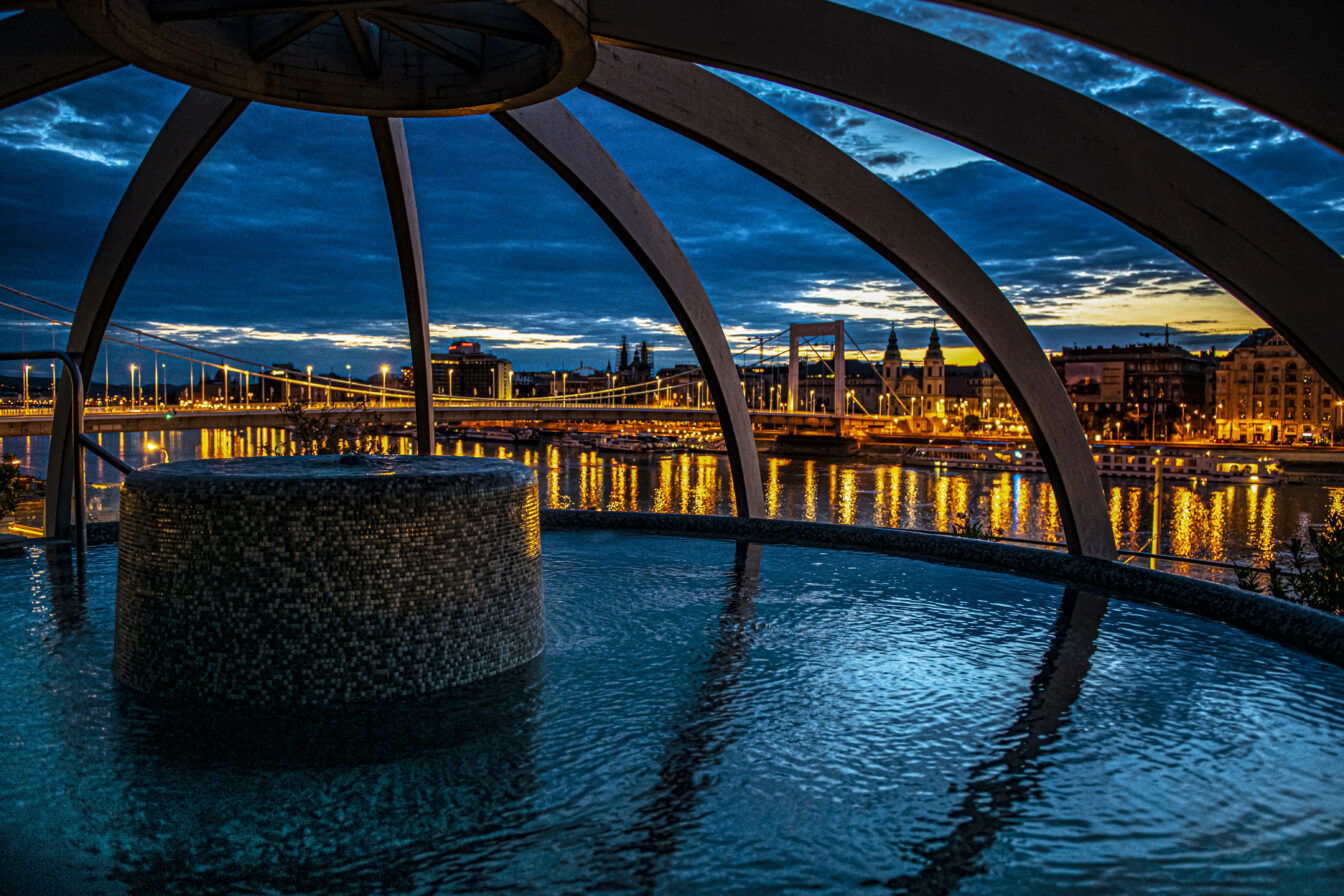 Das Bild zeigt die Rudas Therme in Budapest bei Nacht. Es bietet einen atemberaubenden Blick auf die beleuchtete Elisabethbrücke und die Donau, eingerahmt von einer modernen Kuppelstruktur über dem Thermalbecken. Die Stadtlichter spiegeln sich im Wasser und schaffen eine beruhigende, entspannende Atmosphäre.