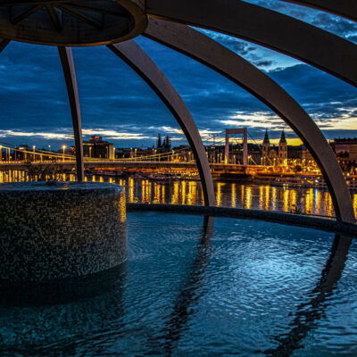 Das Bild zeigt die Rudas Therme in Budapest bei Nacht. Es bietet einen atemberaubenden Blick auf die beleuchtete Elisabethbrücke und die Donau, eingerahmt von einer modernen Kuppelstruktur über dem Thermalbecken. Die Stadtlichter spiegeln sich im Wasser und schaffen eine beruhigende, entspannende Atmosphäre.