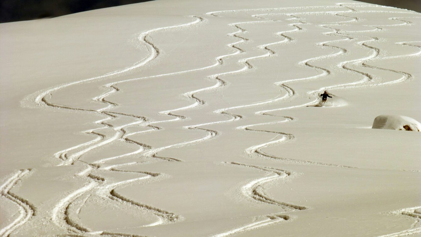 Eine verschneite, unberührte Piste mit zahlreichen eleganten Zickzack-Spuren, die von Skifahrer*innen im Pulverschnee hinterlassen wurden. Eine einzelne Person fährt in der Ferne den Hang hinunter.