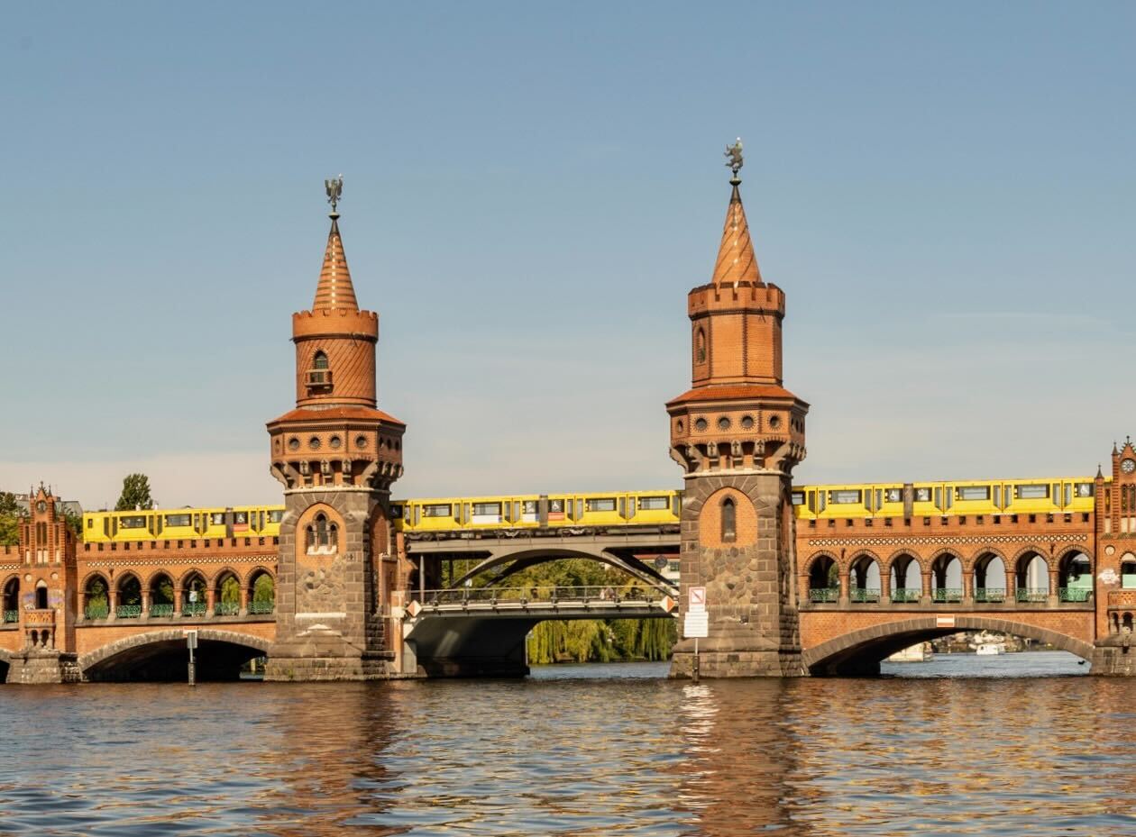 Das Bild zeigt die Oberbaumbrücke in Berlin, eine historische Backsteinbrücke über die Spree mit zwei markanten Türmen. Eine gelbe U-Bahn fährt auf der oberen Ebene der Brücke, während die untere Ebene für Fußgänger und Autos genutzt wird. Im Vordergrund fließt die Spree ruhig unter der Brücke.