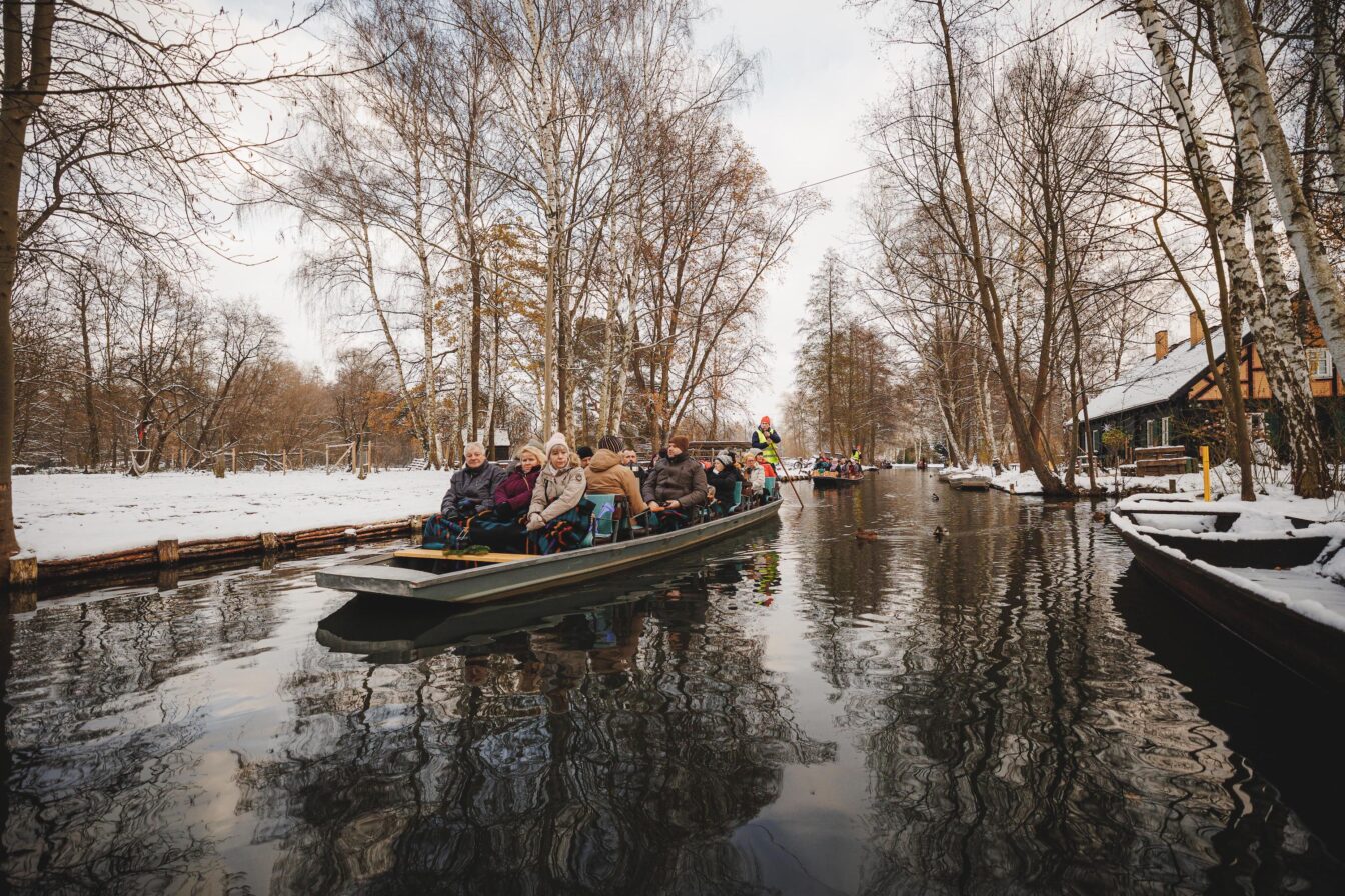 Auf dem Bild ist eine winterliche Szene im Spreewald zu sehen, wo eine Gruppe von Menschen warm eingepackt auf einem traditionellen Kahn durch verschneite, von kahlen Bäumen gesäumte Kanäle fährt. Der Boden und die Ufer sind mit Schnee bedeckt, während der ruhige Fluss das winterliche Umfeld reflektiert. Der Kahn wird von einem Fährmann in bunter Kleidung gesteuert, während sich im Hintergrund weitere Boote auf dem Wasser befinden.