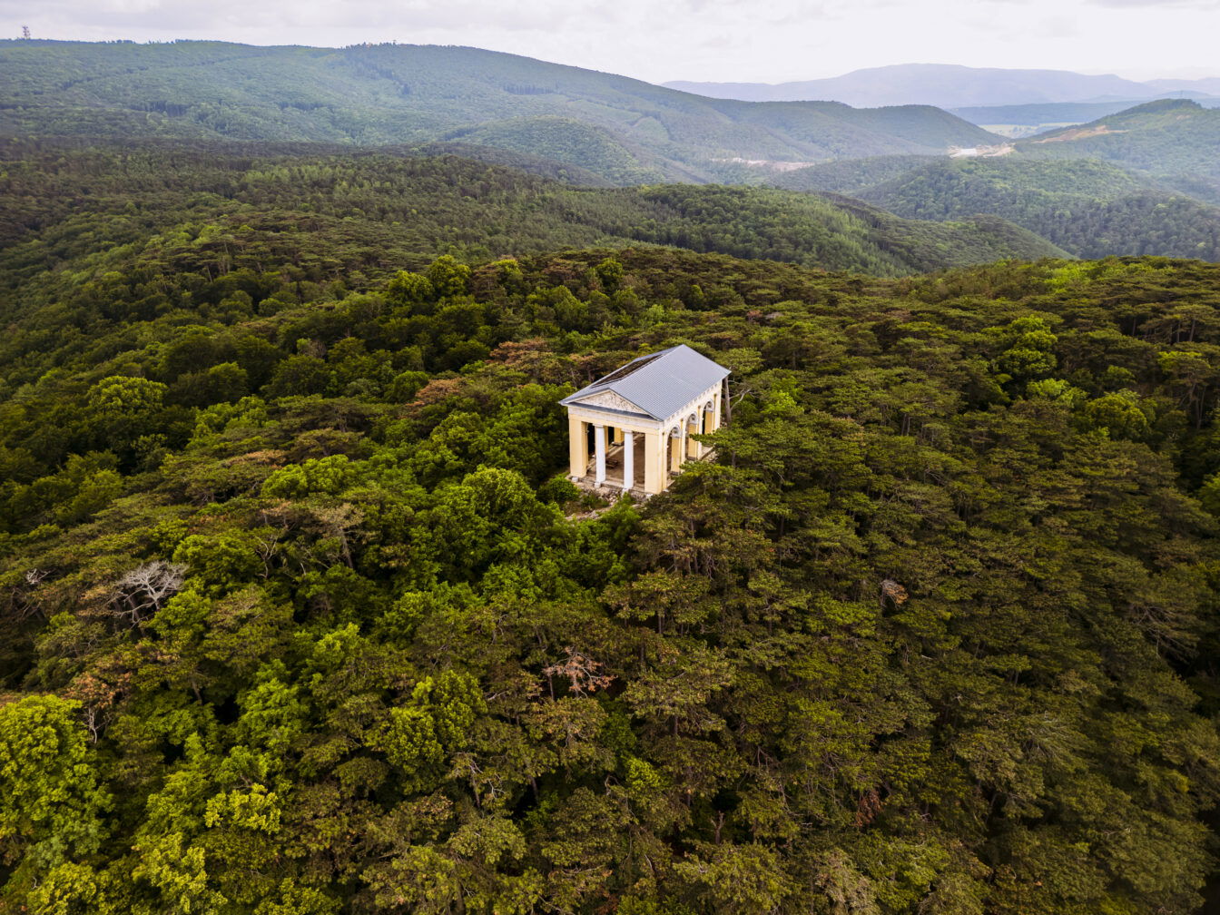 Luftaufnahme des Husarentempels im Wienerwald, der auf einem bewaldeten Hügel liegt. Der Tempel, ein neoklassizistisches Bauwerk mit Säulen und einem Dreiecksgiebel, ist von dichten grünen Bäumen umgeben. Im Hintergrund erstrecken sich bewaldete Hügel und eine weite, hügelige Landschaft. Der Himmel ist leicht bewölkt und verleiht der Szene eine ruhige und idyllische Atmosphäre.