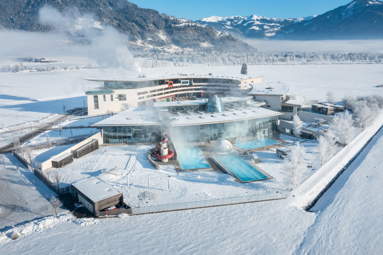 Luftaufnahme des Tauern Spa in Zell am See im Winter. Das moderne Spa-Gebäude mit großen Glasfassaden und einer kurvigen Architektur liegt inmitten einer verschneiten Landschaft, umgeben von schneebedeckten Feldern und Bergen im Hintergrund. Der Dampf steigt von den Außenpools auf, während die Sonne das Gelände in ein helles, kaltes Winterlicht taucht. Die Pools und Wasserattraktionen sind klar sichtbar, mit Rutschen und beheizten Becken, die ein Kontrast zum Schnee darstellen.