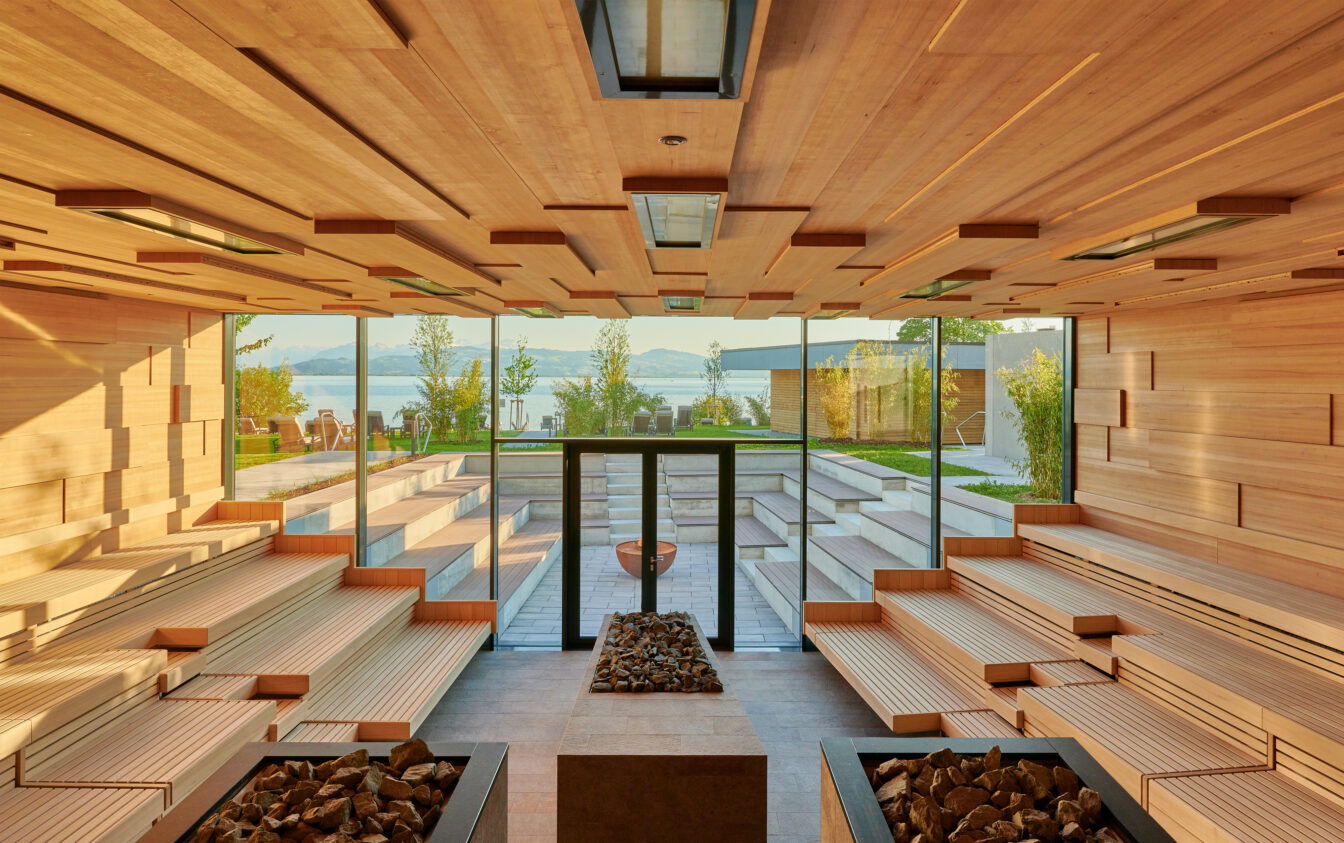 Therme Lindau, Das Bild zeigt einen modernen, großzügigen Sauna-Raum mit einer beeindruckenden Aussicht auf eine Gartenanlage und einen See im Hintergrund. Die Sauna hat ein klares, minimalistisches Design mit hellen Holzbänken und geometrischen Holzverkleidungen an der Decke. Große Fensterfronten lassen viel natürliches Licht herein und eröffnen einen atemberaubenden Blick auf die umliegende Natur und das Wasser, wodurch eine ruhige und entspannende Atmosphäre geschaffen wird.