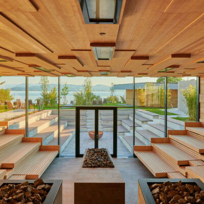 Therme Lindau, Das Bild zeigt einen modernen, großzügigen Sauna-Raum mit einer beeindruckenden Aussicht auf eine Gartenanlage und einen See im Hintergrund. Die Sauna hat ein klares, minimalistisches Design mit hellen Holzbänken und geometrischen Holzverkleidungen an der Decke. Große Fensterfronten lassen viel natürliches Licht herein und eröffnen einen atemberaubenden Blick auf die umliegende Natur und das Wasser, wodurch eine ruhige und entspannende Atmosphäre geschaffen wird.