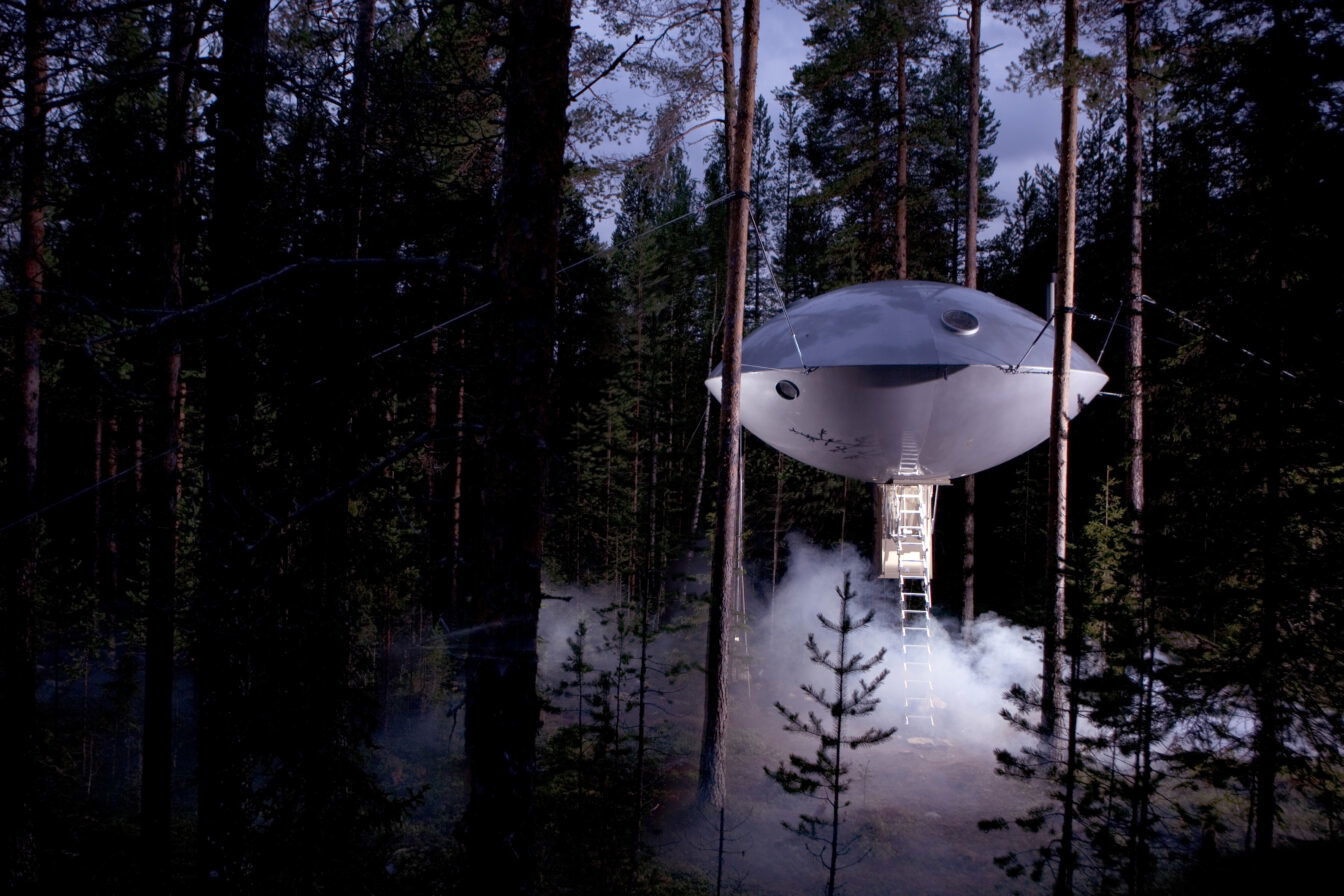 Das Bild zeigt ein futuristisch anmutendes Baumhaus in der Form eines UFOs, das zwischen hohen Bäumen im Wald schwebt. Eine Nebel- oder Rauchschicht umgibt das Baumhaus, was die geheimnisvolle Atmosphäre verstärkt. Eine Leiter führt von der Bodenebene zur Öffnung des UFO-artigen Baumhauses hinauf. Die Szenerie wirkt wie aus einem Science-Fiction-Film, eingebettet in eine natürliche Waldlandschaft.