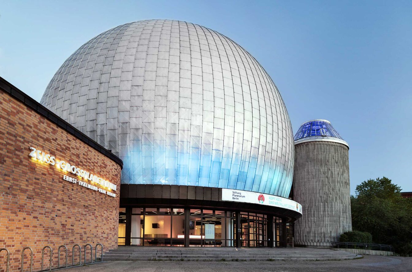 Außenansicht des Zeiss-Großplanetariums in Berlin. Die große, silberne Kuppel des Planetariums dominiert das Bild, daneben steht ein weiteres, kleineres Gebäude mit einer blauen Glaskuppel. Links im Bild ist ein Teil des Backsteingebäudes mit einem beleuchteten Schild zu sehen, auf dem 'Zeiss-Großplanetarium Ernst-Thälmann-Park Berlin' steht.