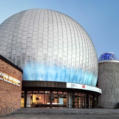 Außenansicht des Zeiss-Großplanetariums in Berlin. Die große, silberne Kuppel des Planetariums dominiert das Bild, daneben steht ein weiteres, kleineres Gebäude mit einer blauen Glaskuppel. Links im Bild ist ein Teil des Backsteingebäudes mit einem beleuchteten Schild zu sehen, auf dem 'Zeiss-Großplanetarium Ernst-Thälmann-Park Berlin' steht.