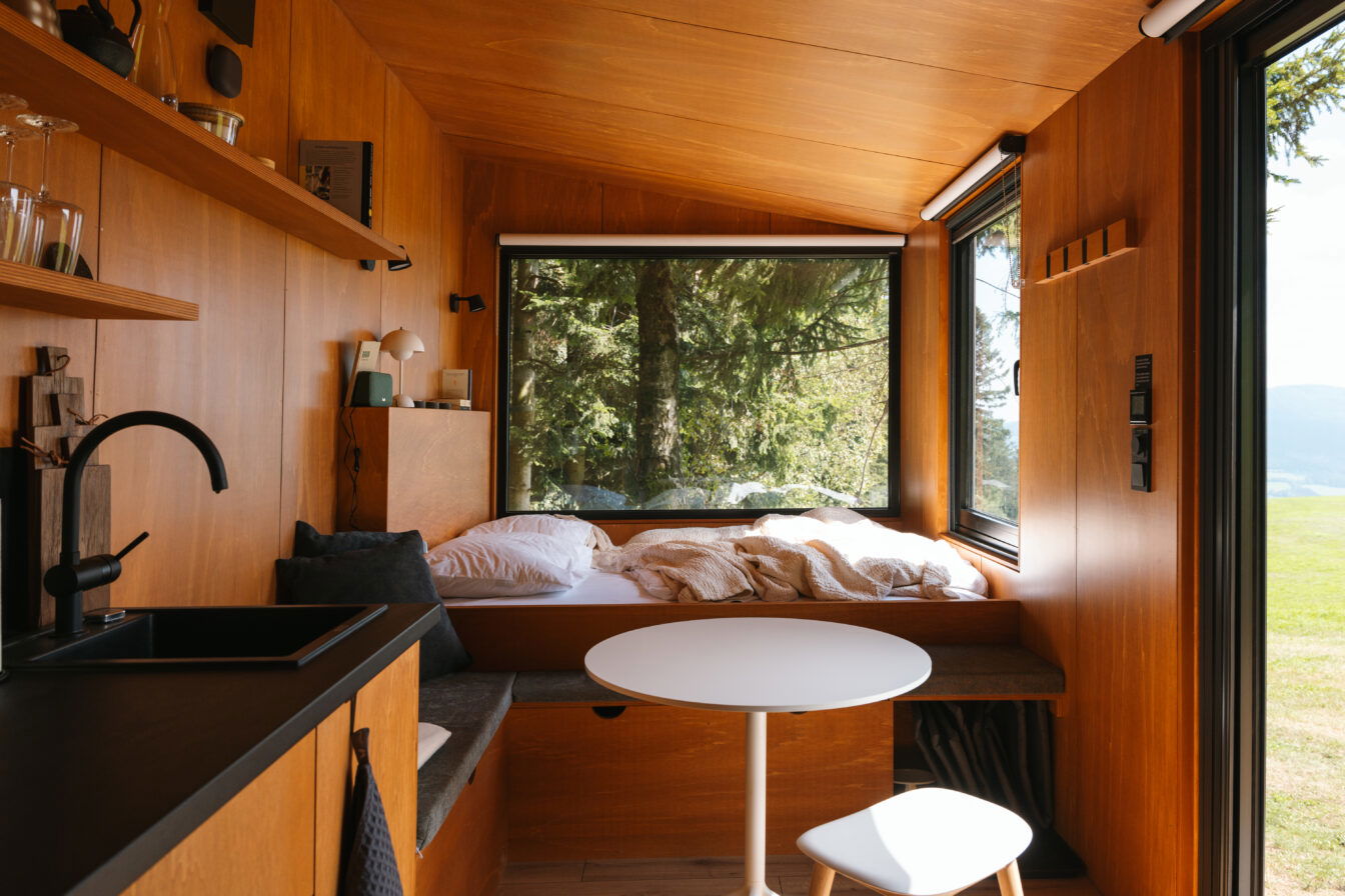 Ein gemütliches Tiny House aus Holz mit einem großen Panoramafenster, das den Blick auf eine grüne Waldlandschaft freigibt. Links befindet sich eine kleine Küchenzeile mit schwarzem Wasserhahn und Regalen. Im Hintergrund liegt ein Bett unter dem Fenster, mit weißen und beigen Decken und Kissen.